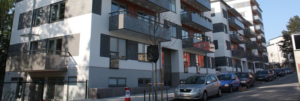 Vitt lägenhetshus med grå detaljer, som har balkonger som vetter mot en gata där bilar står parkerade.