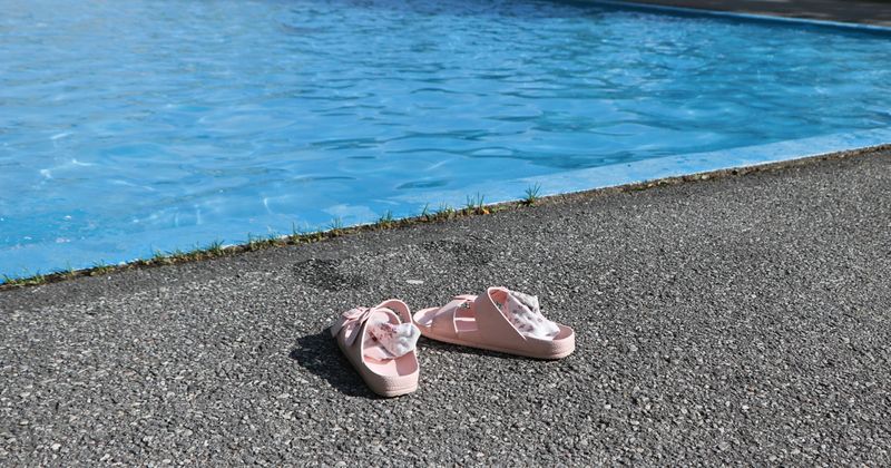 Två rosa sandaler med ljusa strumpor nerknycklat i dem står vid kanten av en plaskdamm. På långt håll ser man ett barn som badar.