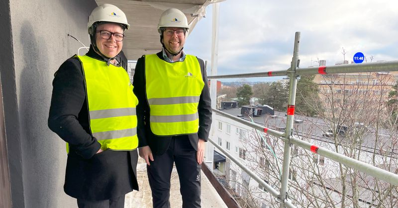 Två män i gula varselvästar och vita bygghjälmar på en balkong med byggnadsställning utanför.