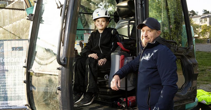 En flicka sitter i en grävmaskin och en man står utanför hytten, båda ler mot kameran.