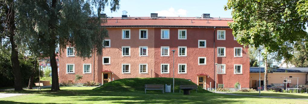Ett lägenhetshus i röda nyanser med två ingångar. Framför huset finns en grön kulle och träd.