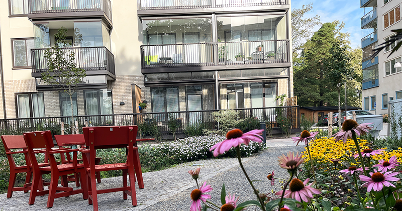 Uinnergård med ett rött bord och röda trästolar. Rosa och gula blommor i förgrunden. Hus i bakgrunden med vit fasad.