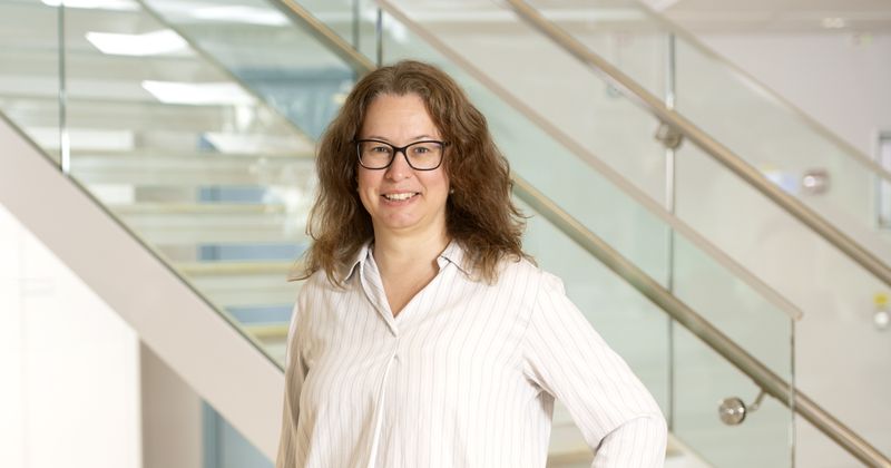 Bild på kvinna som står inomhus framför en trappa iklädd vid skjorta med beigea ränder diagonalt, med glasögon och brunt, lite lockigt, långt hår.