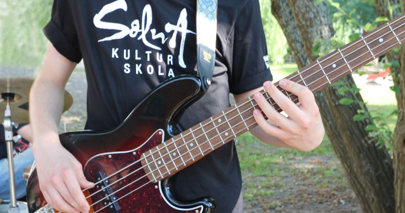Ung pojke som spelar gitarr och med en svart t-shirt på sig med Solna Kulturskolas logga på bröstet. Utomhus och det är soligt.