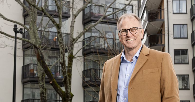 Man med brun rock, glasögon med svarta bågar och ljusblå skjorta framför husfasad. Fasaden har balkonger och mellan hus och man står ett träd som man ser delar av.