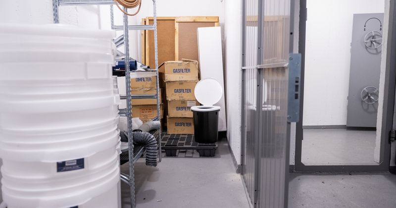 Bilden visar ett lager inuti ett skyddsrum. I förgrunden ser man vita plasthinkar och längst in står en uppfälld engångstoalett samt ett antal kartoner.