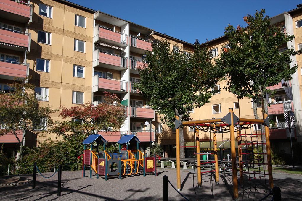 Gult lägenhetshus med 5 våningar, röda balkonger. I förgrunden en lekplats på innergården.