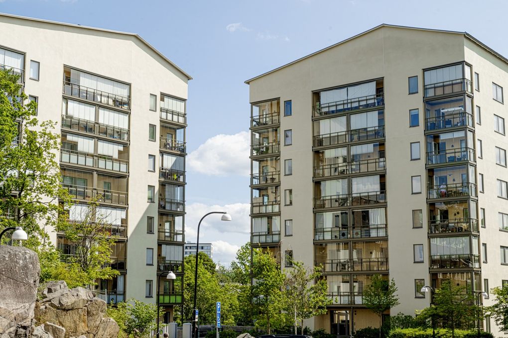 Två vita flervåningshus med inglasade balkonger, omgivna av grönska.