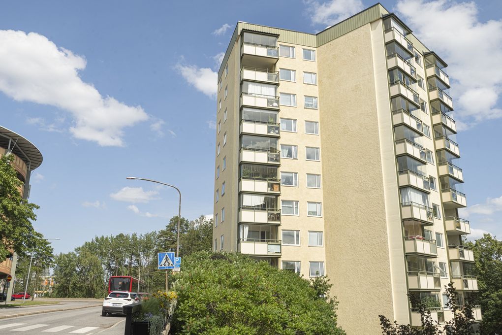 Ljust höghus med balkonger vid en gata där bilar står parkerade.