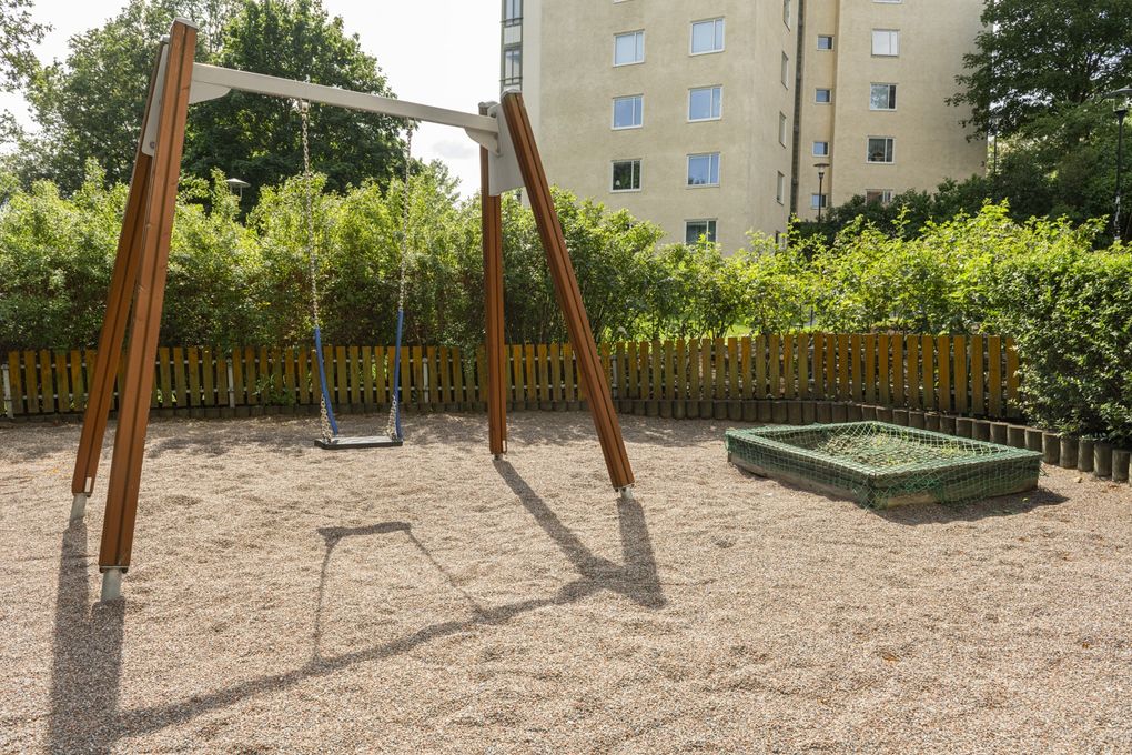 Lekplats med gunga och sandlåda omgivet av grönska, med ett gult höghus i bakgrunden.