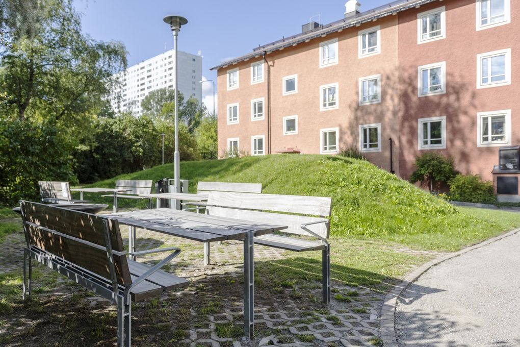 Bord och bänkar vid en grönyta med ett rött flerbostadshus i bakgrunden.