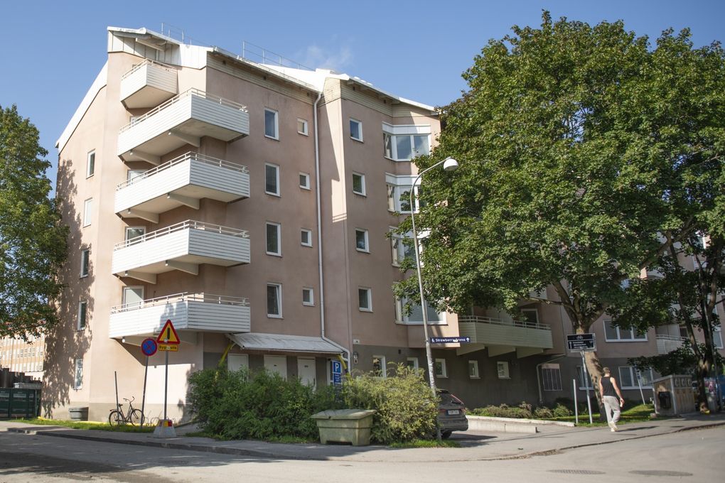 Rosa flerbostadshus med vita balkongen vid en gata. Ett stort grönskande träd står framför huset.