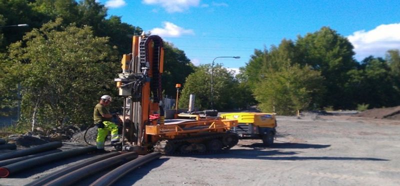 En person i arbetskläder står i soligt väder och utför en geoteknisk undersökning med hjälp av en borrbandvagn.