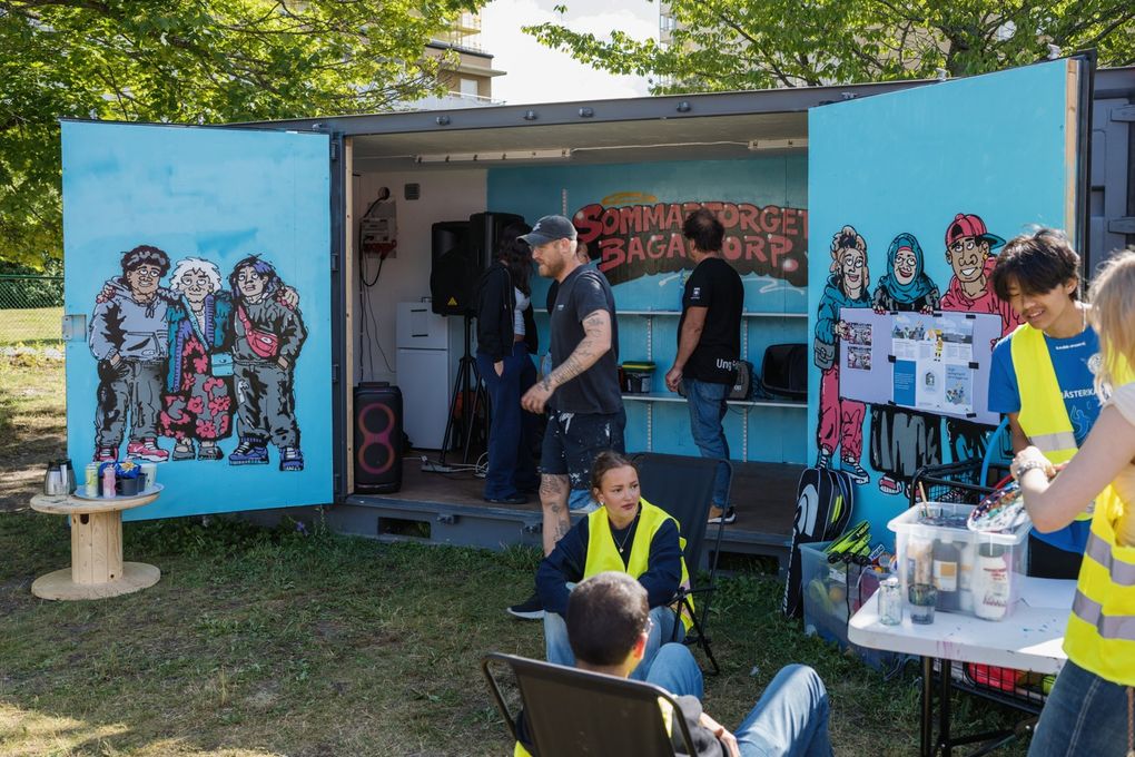 Personer som sitter på stolar i gräset framför en målad container.