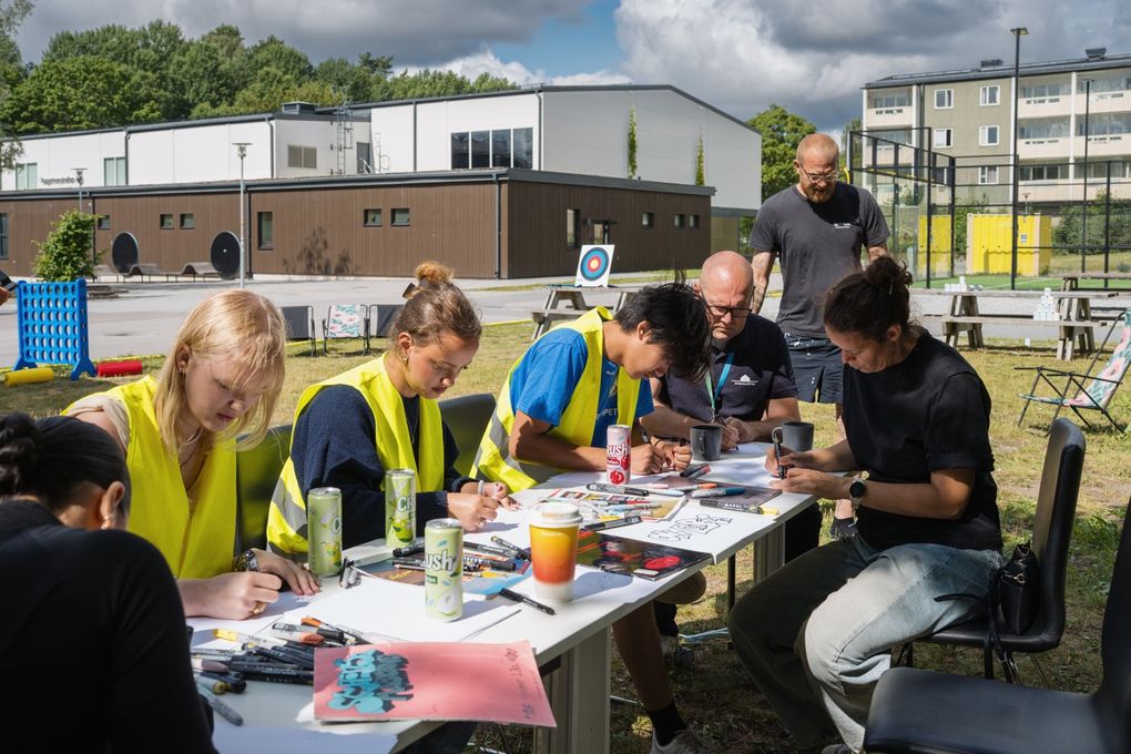 Personer som sitter vid ett bord och målar utomhus.