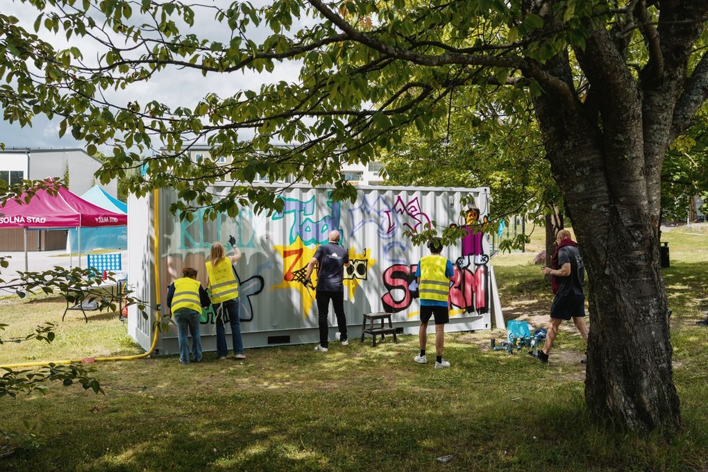 Personer står i gräset och målar graffiti på en vägg.