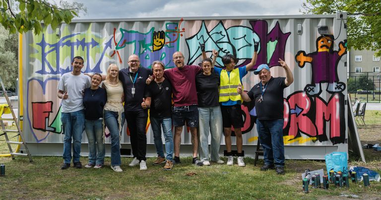 Nio personer står i gräset framför en vägg målad med graffiti.