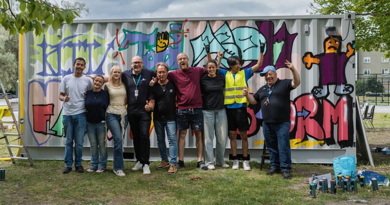 Nio personer står i gräset framför en vägg målad med graffiti.